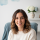 Headshot of a smiling Spanish woman in her late 20s, with brown hair, in front of a subtly blurred background with white and 